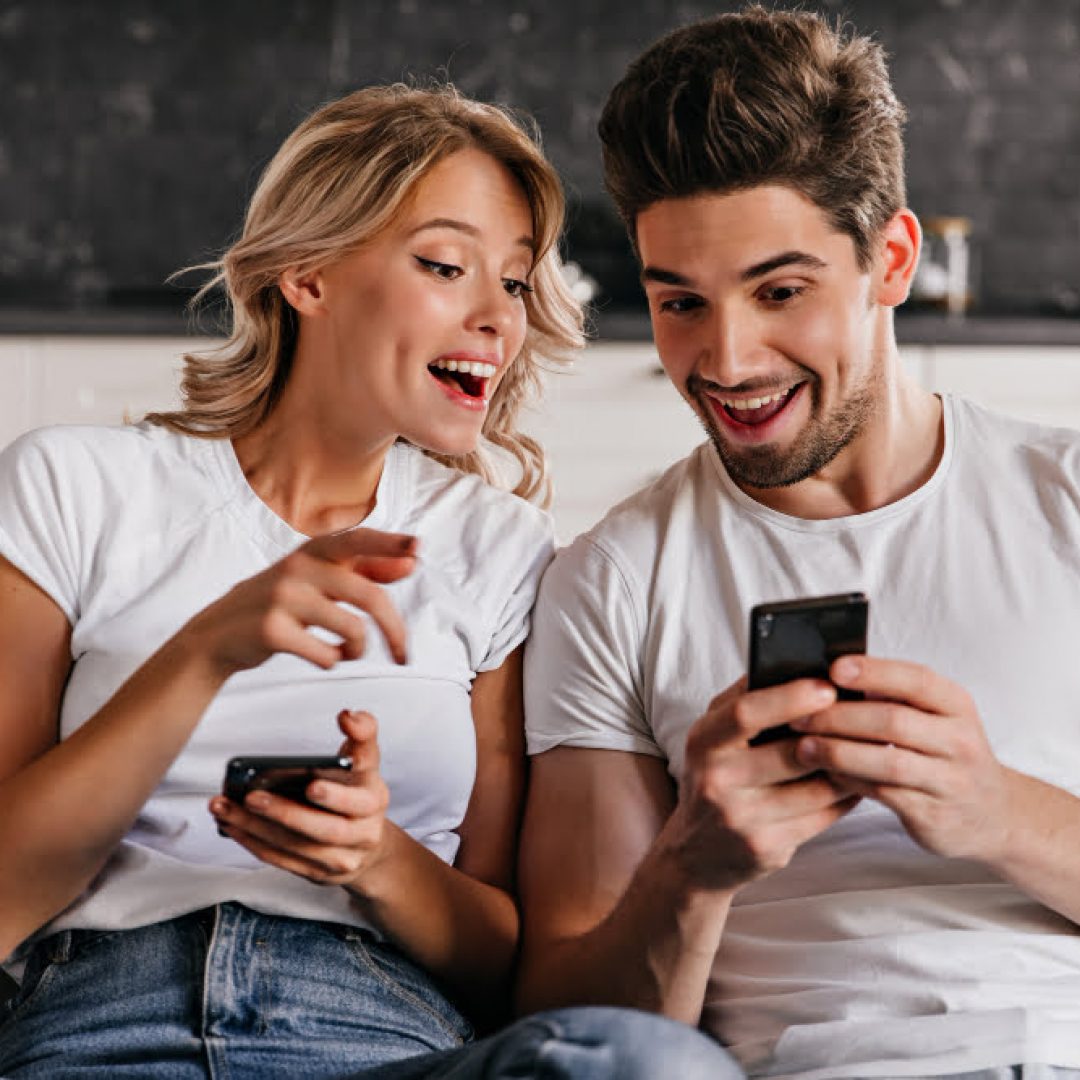 smiling-couple-sitting-couch-with-phones-adorable-young-woman-holding-smartphone
